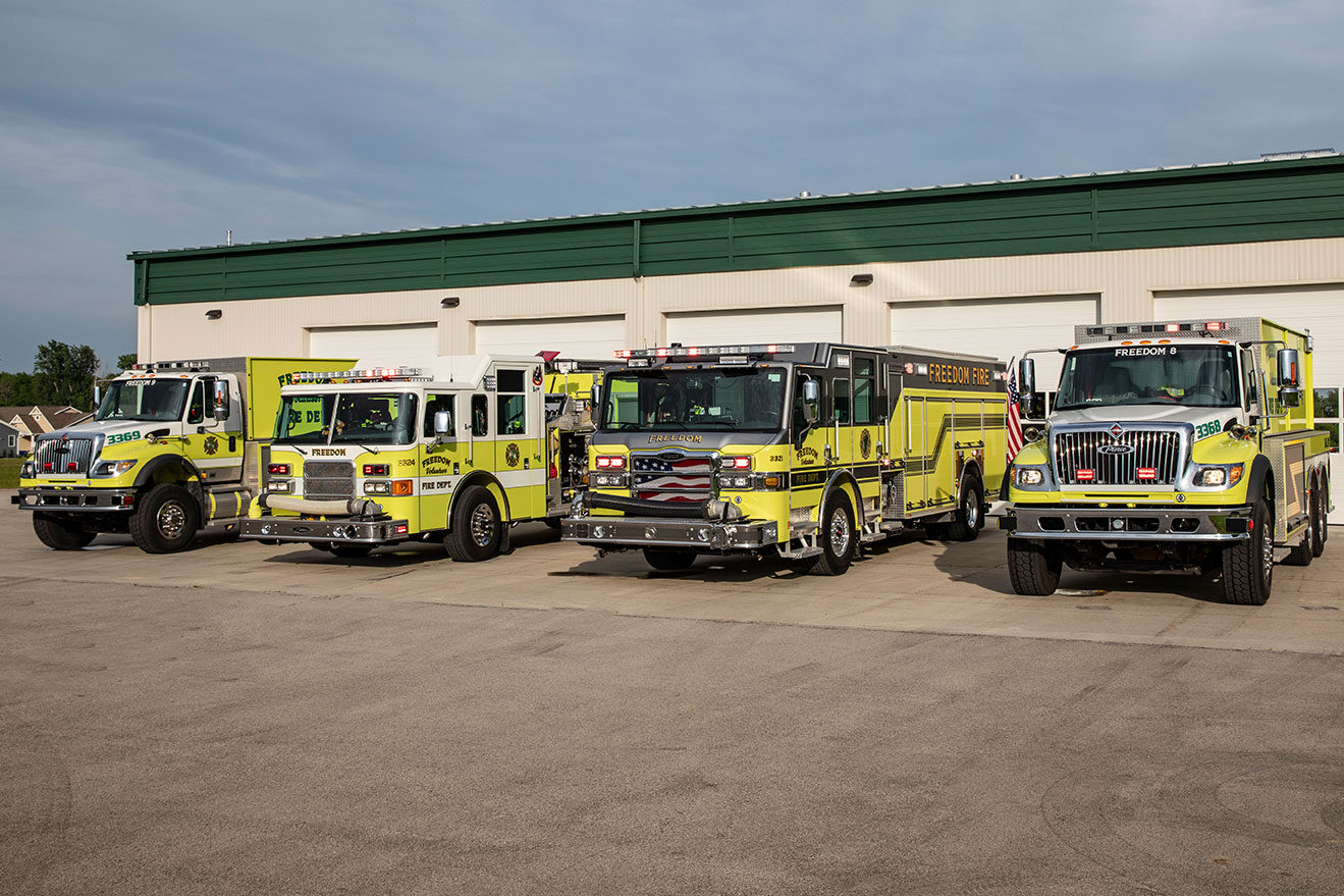 Four fire trucks parked in front of a fire station on a cloudy day with their lights on.