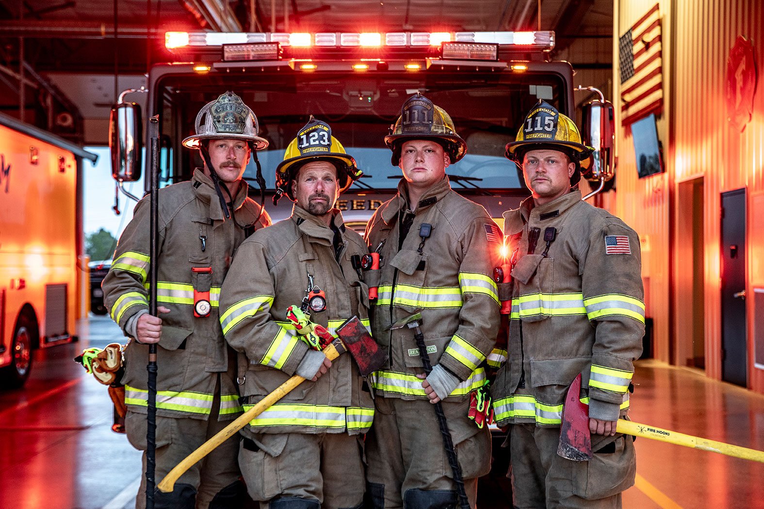 Four firefighters in turnout gear holding firefighting tools standing in front of a fire truck with the lights on at a fire station.