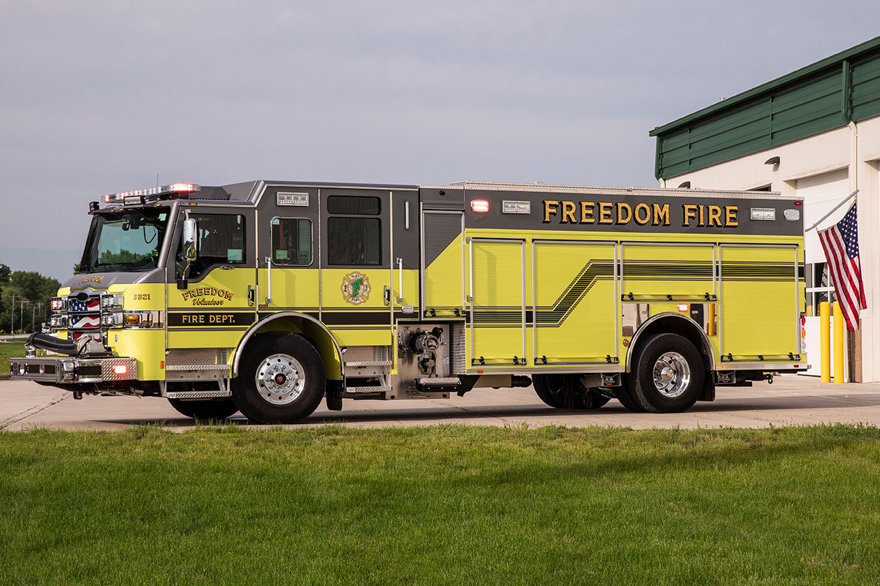 The driver's side of a fire truck with the lights on and an American flag hanging off the rear in front of a fire station.