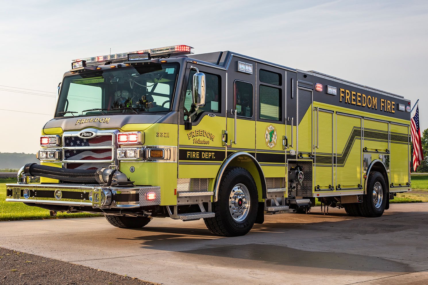 The front driver's side of a yellow fire truck with the lights on and an American flag hanging off the rear.