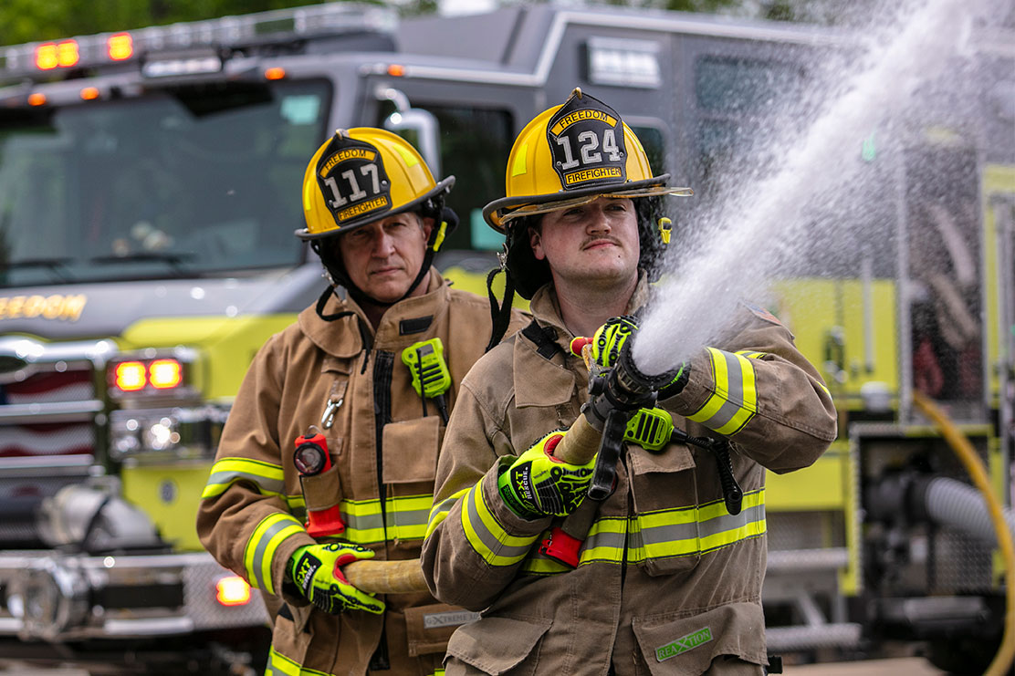 Two firefighters in turnout gear standing in front of a yellow fire truck with the lights on spraying water from a fire hose.