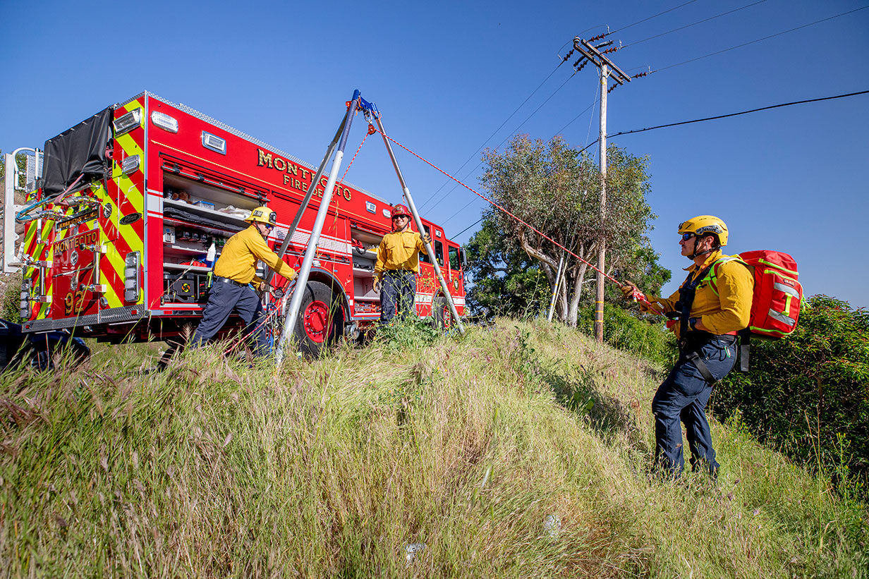 A firefighter repelling down a grass hill from a tripod and two other firefighters operating the rope next to a fire truck.