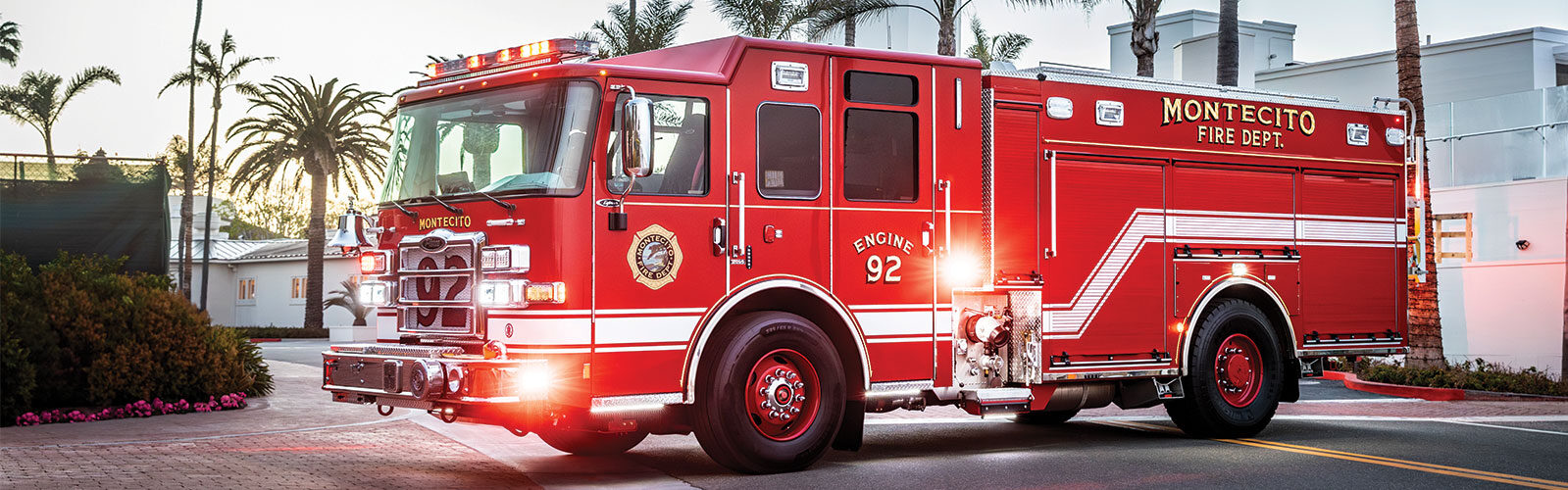 The front driver's side of an Enforcer PUC Pumper parked on a road with the lights on in front of palm trees.