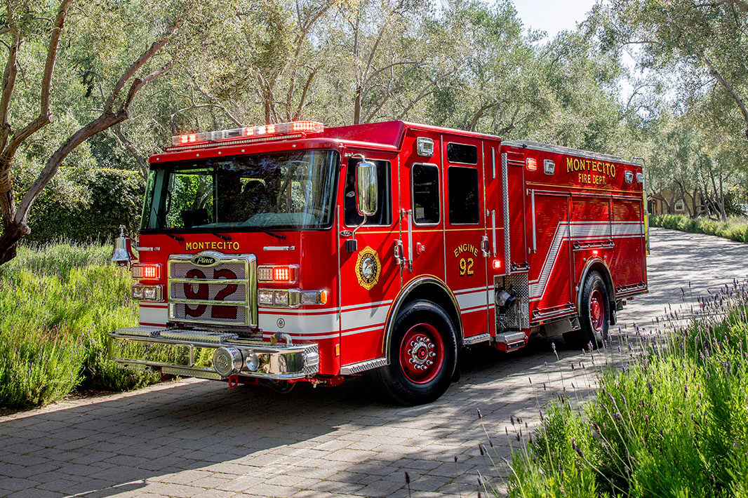 The front driver's side of a fire truck driving down a narrow paved road by green trees and grass.
