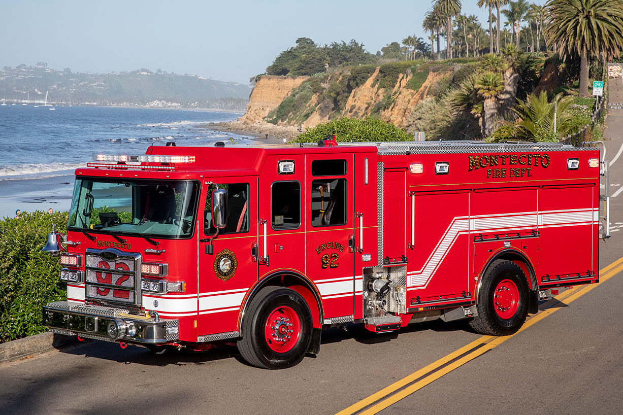 The front driver's side of a fire truck parked on a paved two lane road next to an ocean cliff side.