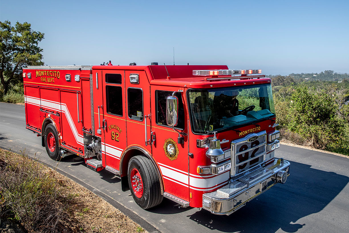 The front officer's side of a fire truck driving down a paved road next to a valley full of green trees.