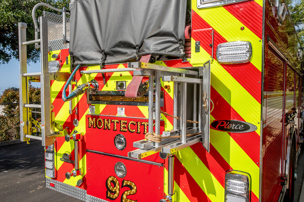 The rear of a fire truck with a compartment open showing ground ladders and a hosebed filled with hose and covered by a black cover.