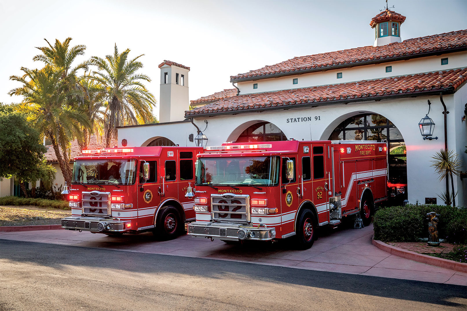Two fire trucks side by side with their lights on pulling out of a fire station next to palm trees on a sunny day.