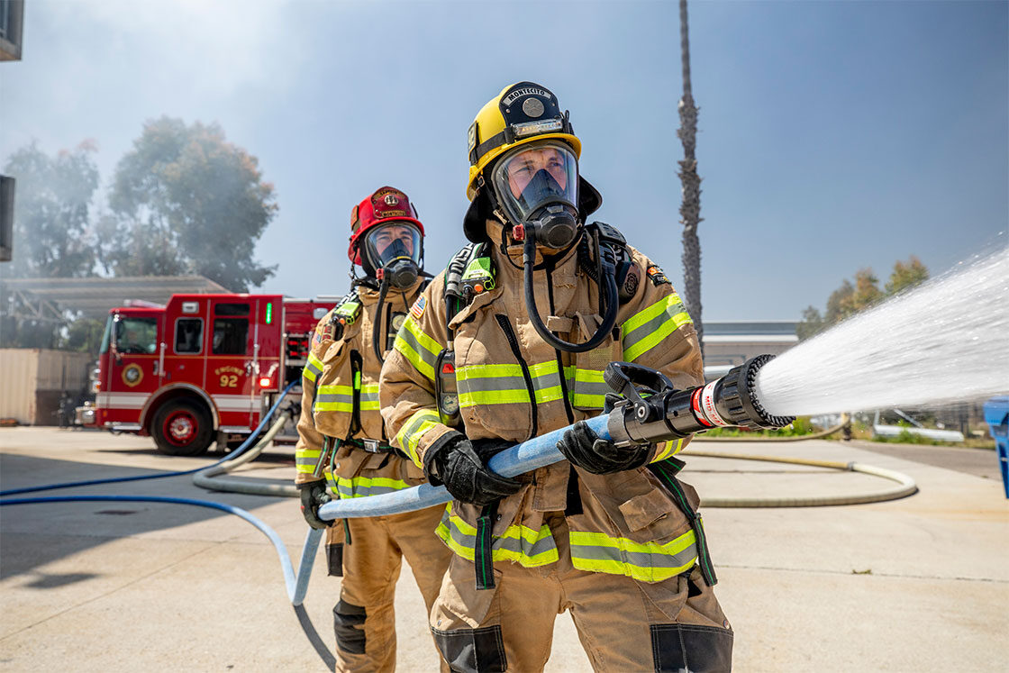 Two firefighters in turnout gear holding on to a firehose spraying water with a fire truck in the background.