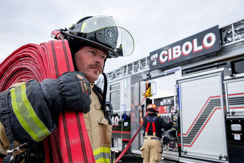 A firefighter in turnout gear carrying hose on his shoulder from a fire truck while another firefighter operates the pump panel.