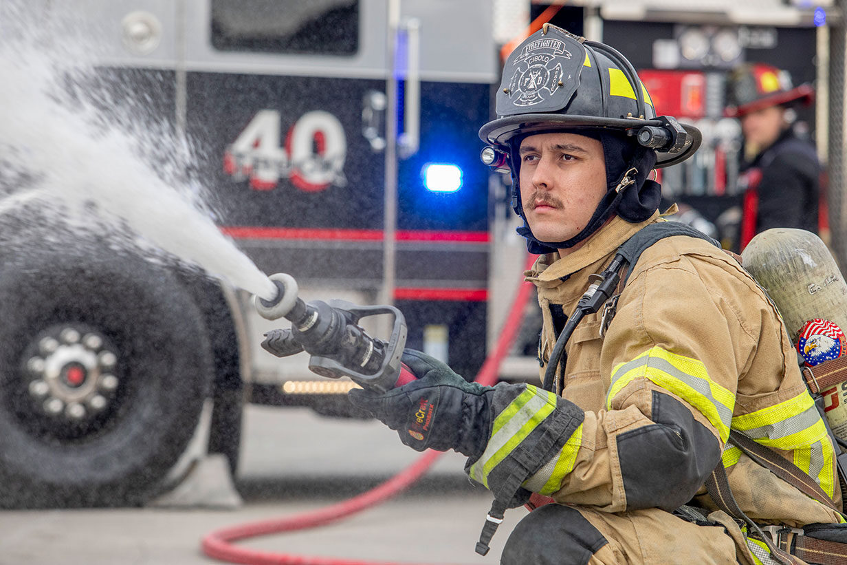 A firefighter in turnout gear kneeling down spraying water from a fire hose while another firefighter is at the pump panel.