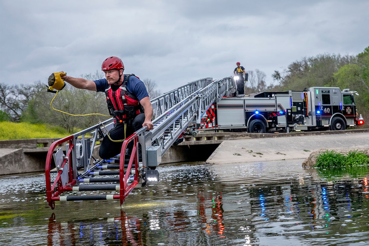A firefighter throwing a rope while sitting on the end of a fire trucks aerial ladder hovering over water.