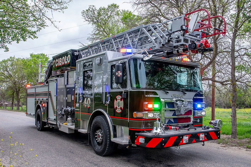 An Enforcer 107 Ascendant Aerial Ladder driving down a paved road in a rural area with the lights on.