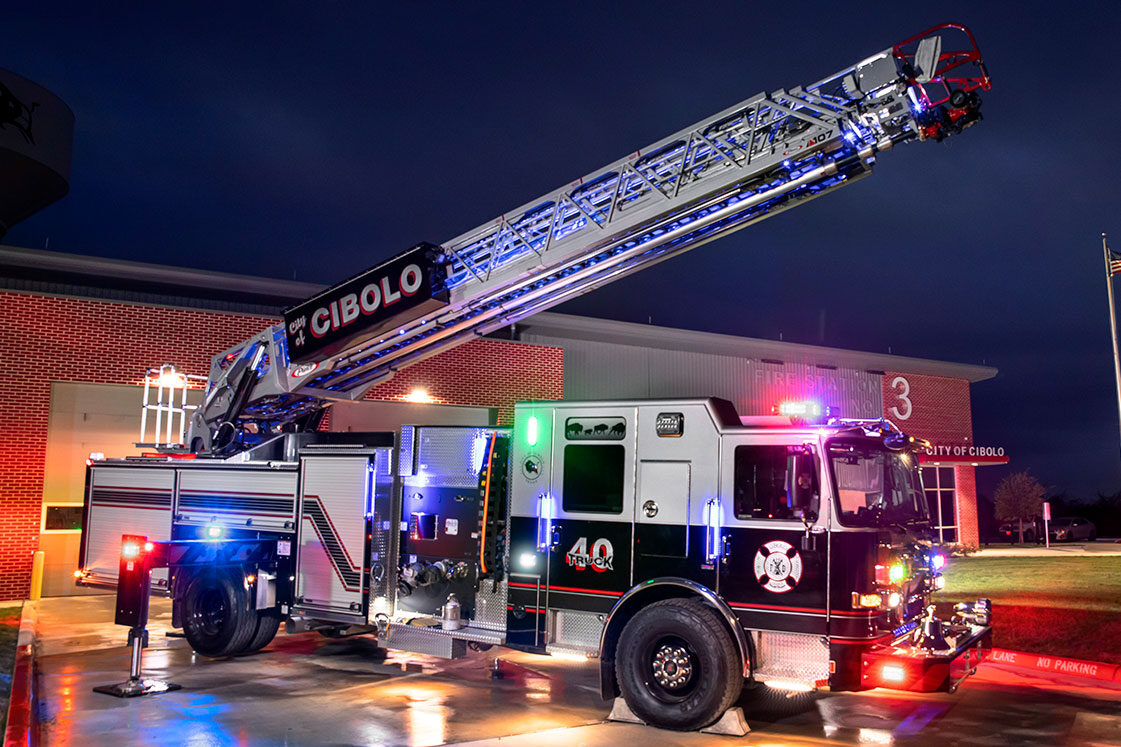 An Enforcer 107 Ascendant Aerial Ladder with the ladder extended and lights on in front of a fire station at night.