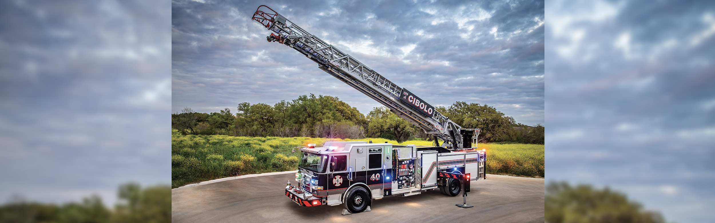 An Enforcer Ascendant 107' Aerial Ladder with the aerial extended and lights on in front of a green grass field on a cloudy day.