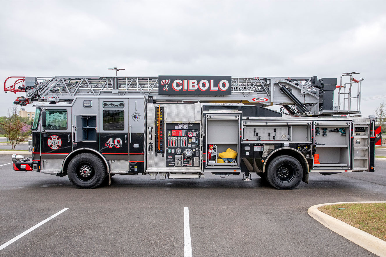 The drivers side of an aerial ladder fire truck with the compartments open showing tools and equipment inside.