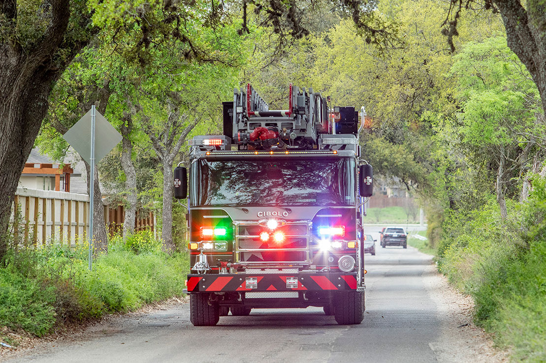 The front of an aerial fire truck with the lights on  driving down a paved road lined with trees.