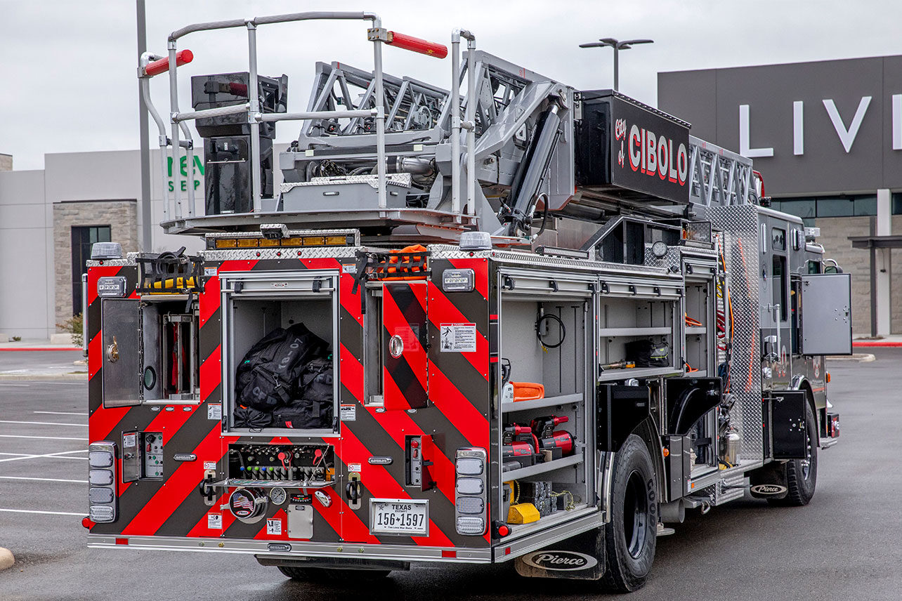 The officers rear side of an aerial ladder fire truck with the compartments open showing ladders, equipment and tools inside.