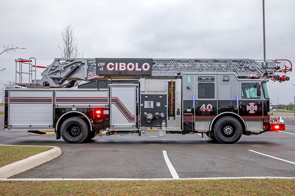 The officers side of an aerial ladder fire truck with the lights on parked in a parking lot on a cloudy day.