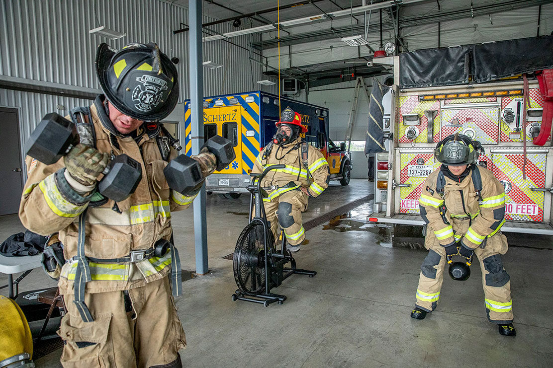 Three firefighters in turnout gear working out with weights in a fire station next to a fire truck and ambulance at a fire station.