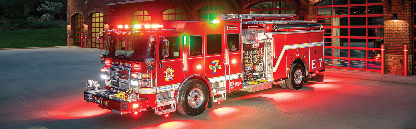The front driver's side of a fire truck with the lights on parked in front of a fire station at night.