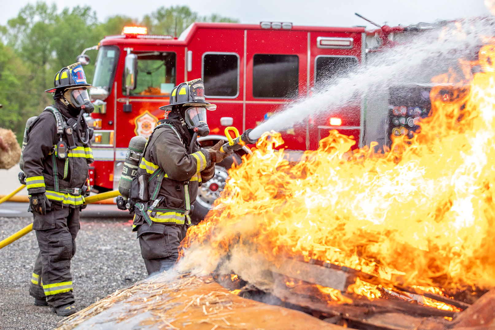 Firefighters in turnout gear spraying water from a fire hose in front of a fire truck on a car fire.