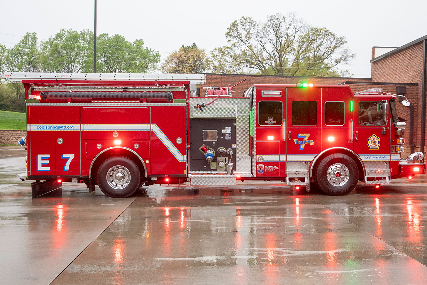 The officers side of an Enforcer pumper with the lights on in a parking lot in front of a fire station on a rainy day.