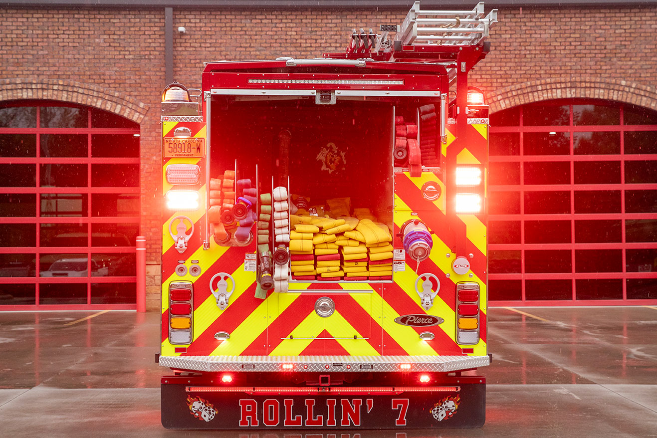 The rear of a fire truck in front of a fire station with the lights on with a hydraulic ladder rack and a hosebed with fire hose.