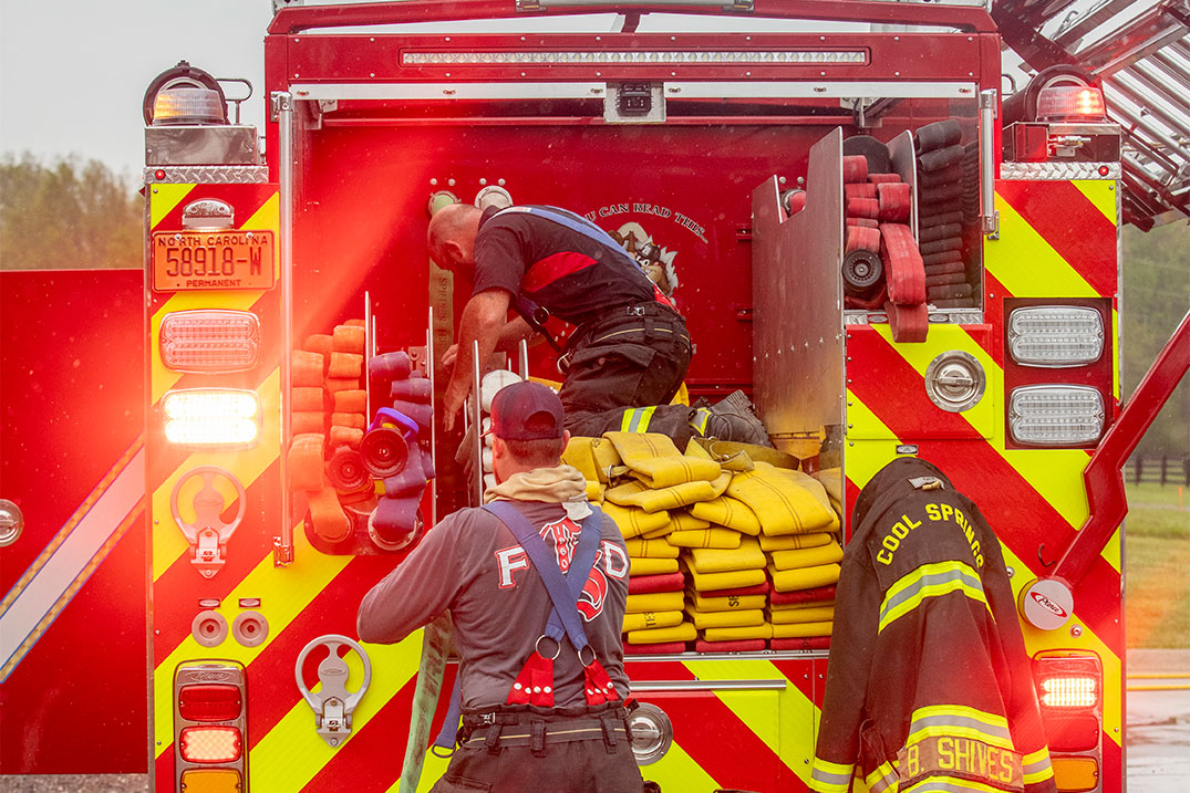 The rear of a fire truck with two firefighters laying fire hose in the hosebed on a rainy day.