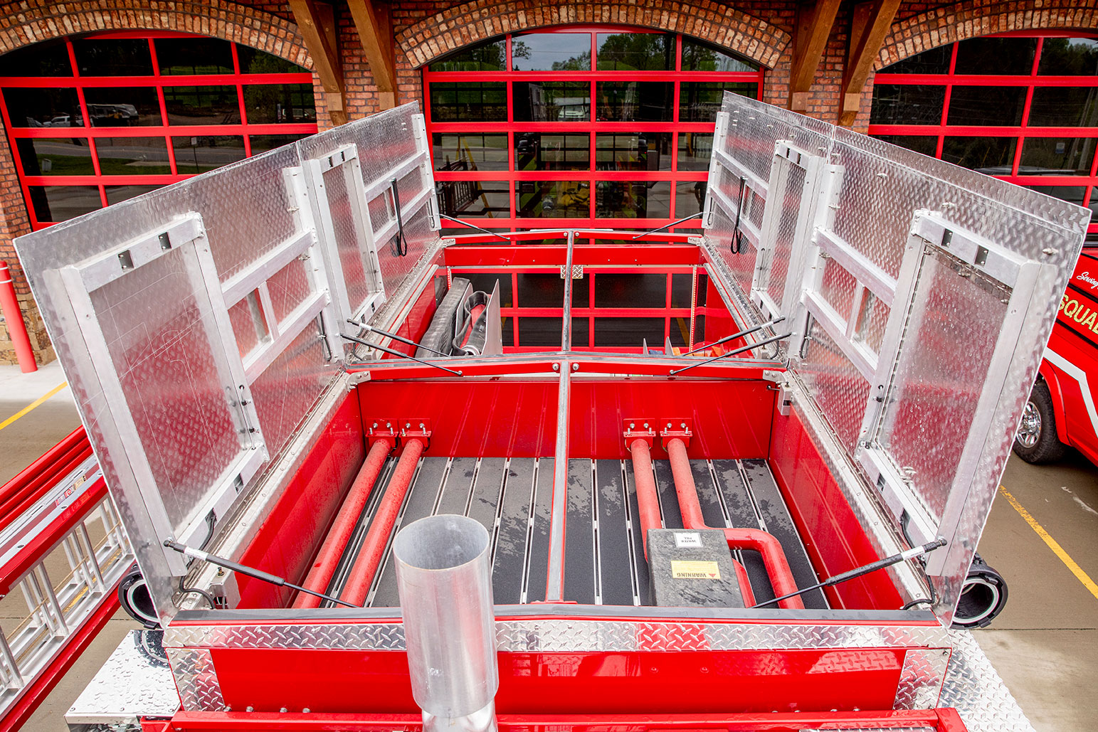 The top of a fire truck with the coffin compartments open in front of a fire station with red glass doors.