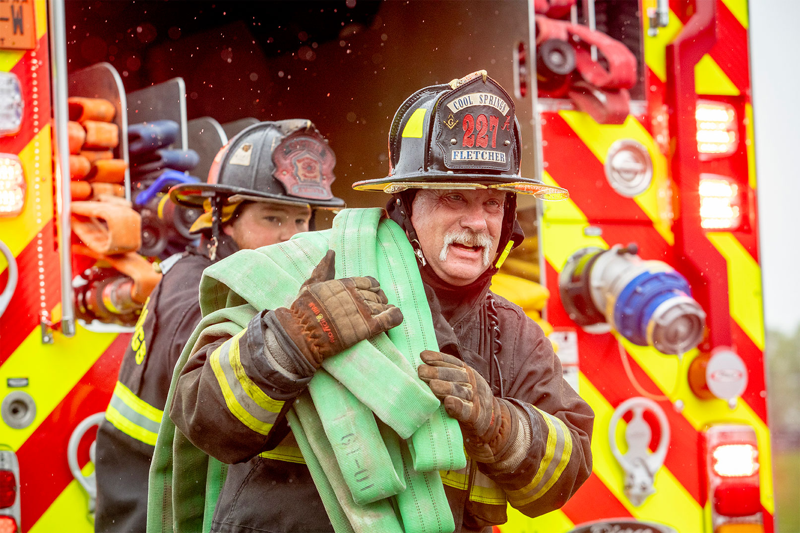 Two firefighters in turnout gear pulling hose off the rear of a fire truck with embers in the air.