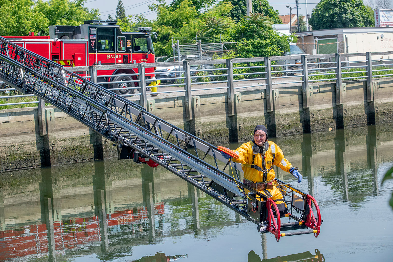 A firefighter in a wet suit on the tip of a fire trucks aerial ladder over water throwing a rope.