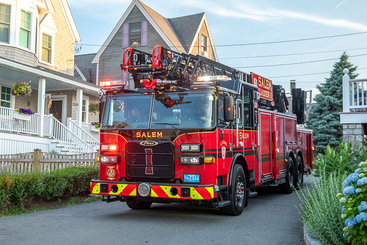 The front drivers side of a 100 Heavy-Duty Low Profile Aerial Ladder driving through a coastal neighborhood with floral landscaping.