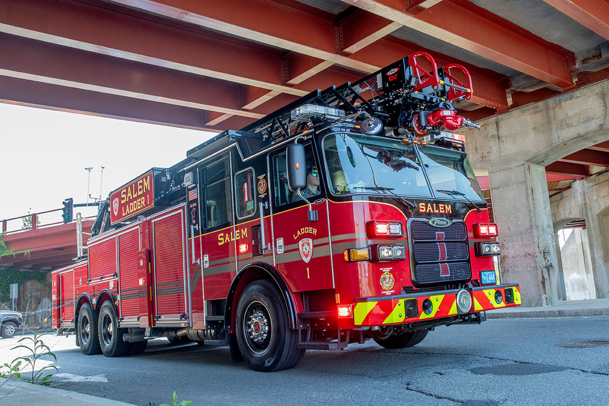 The front officers side of a 100 Heavy-Duty Low Profile Steel Aerial Ladder driving under a bridge with the lights on.