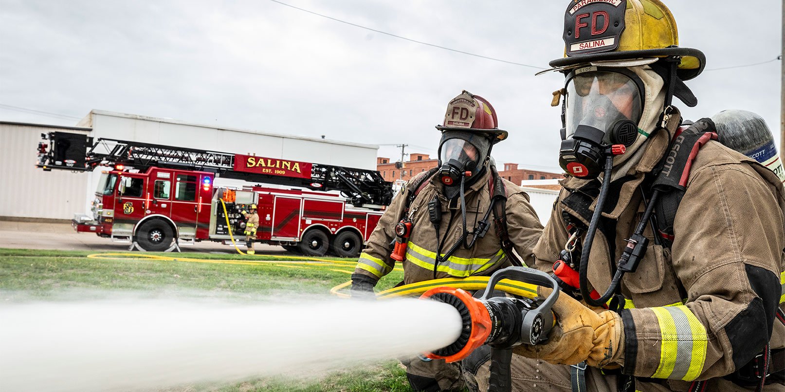 Two firefighters in turnout gear spraying water from a fire hose from a fire truck with a firefighter operating the pump.