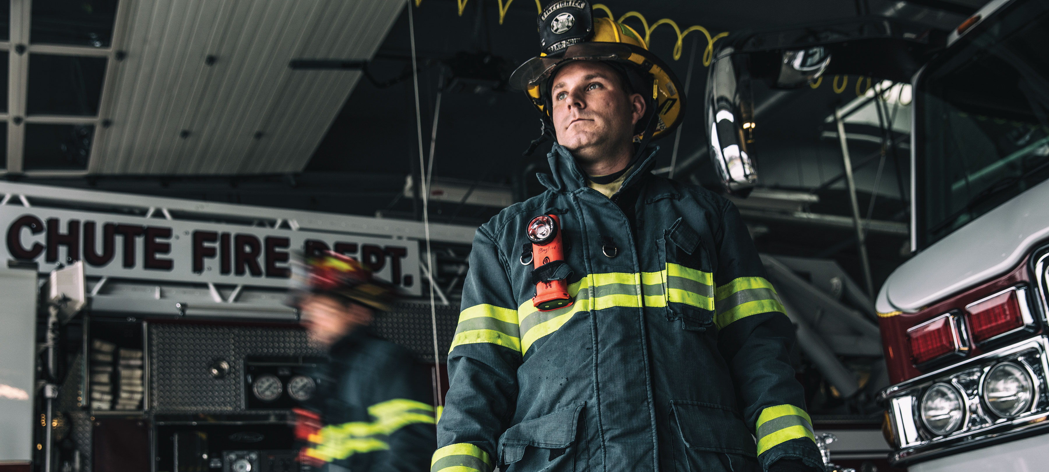 A firefighter in turnout gear standing in the garage of a fire station near an apparatus.