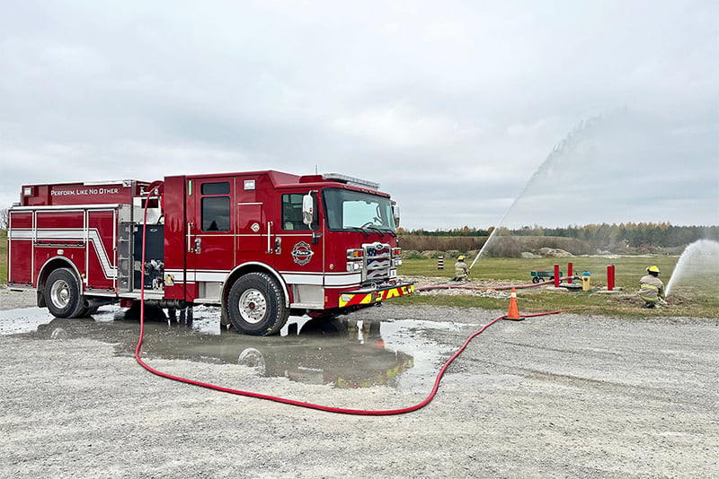 A red Pierce Volterra EV is parked in a gravel lot with a red fire hose extending out of the apparatus on either side reaching several firefighters spraying water in a field.