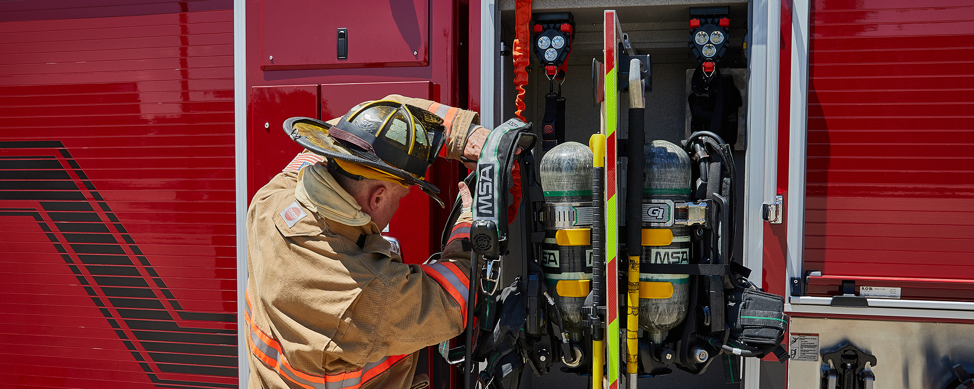 Firefighter putting SCBA and turnout gear onto a clean cab pull out tool board in the side storage compartment on a Pierce fire truck. 