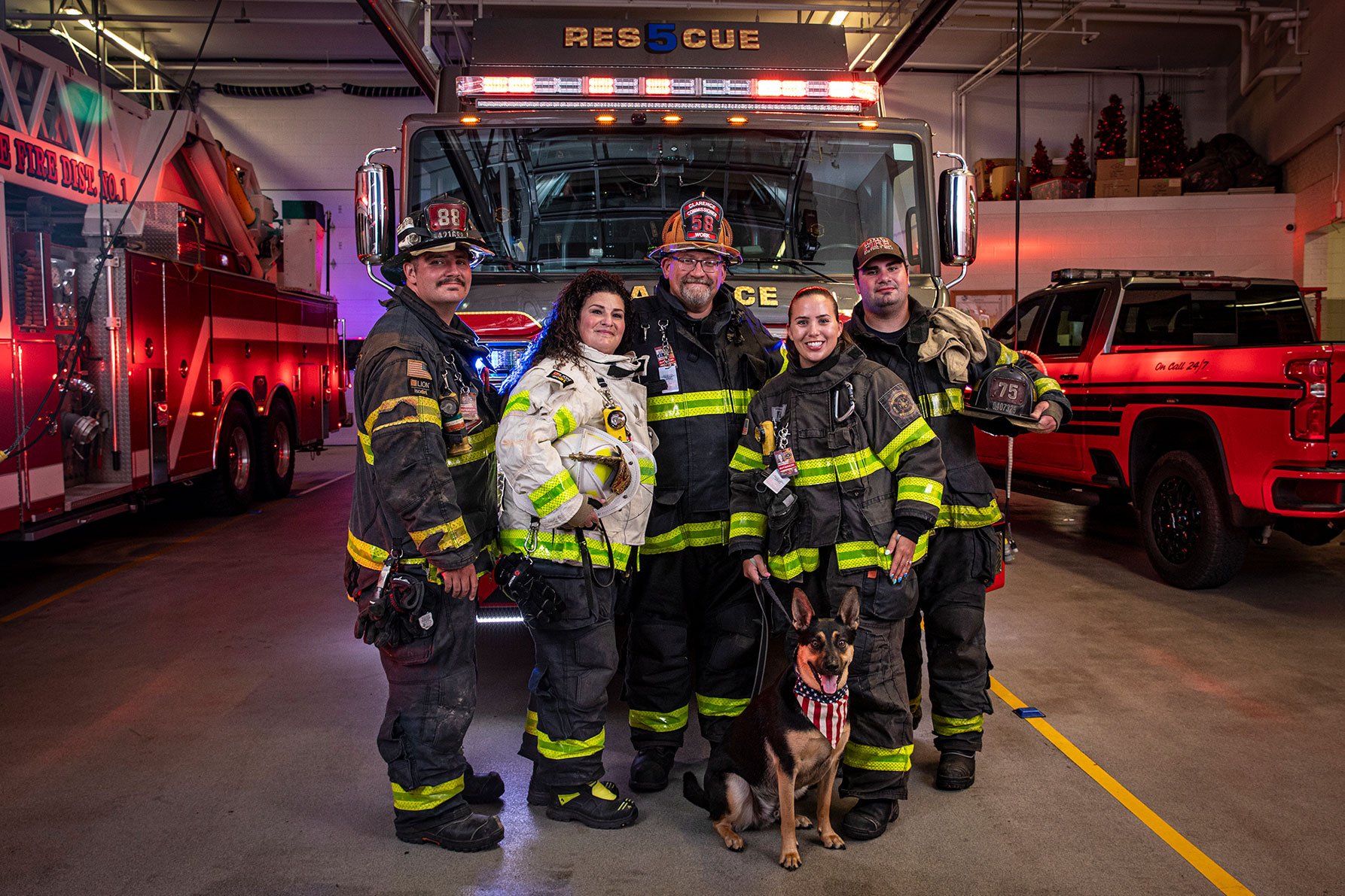 A group of firefighters in turnout gear and a dog standing in front of a HDR fire truck with the lights on in a fire station.