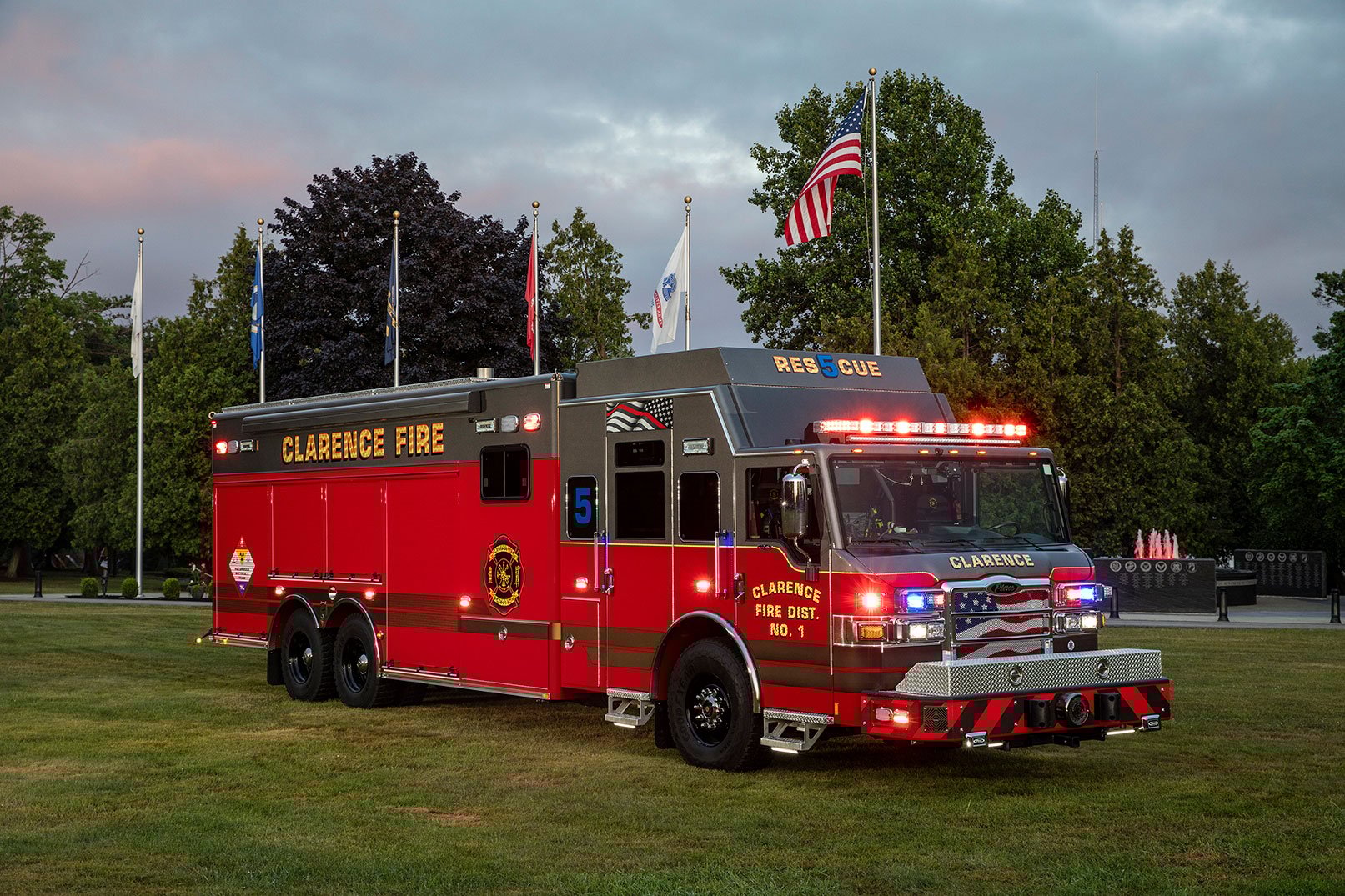 The front officer's side of a Velocity Combination HDR fire truck with the lights on parked in grass beside flagpoles on a cloudy day.