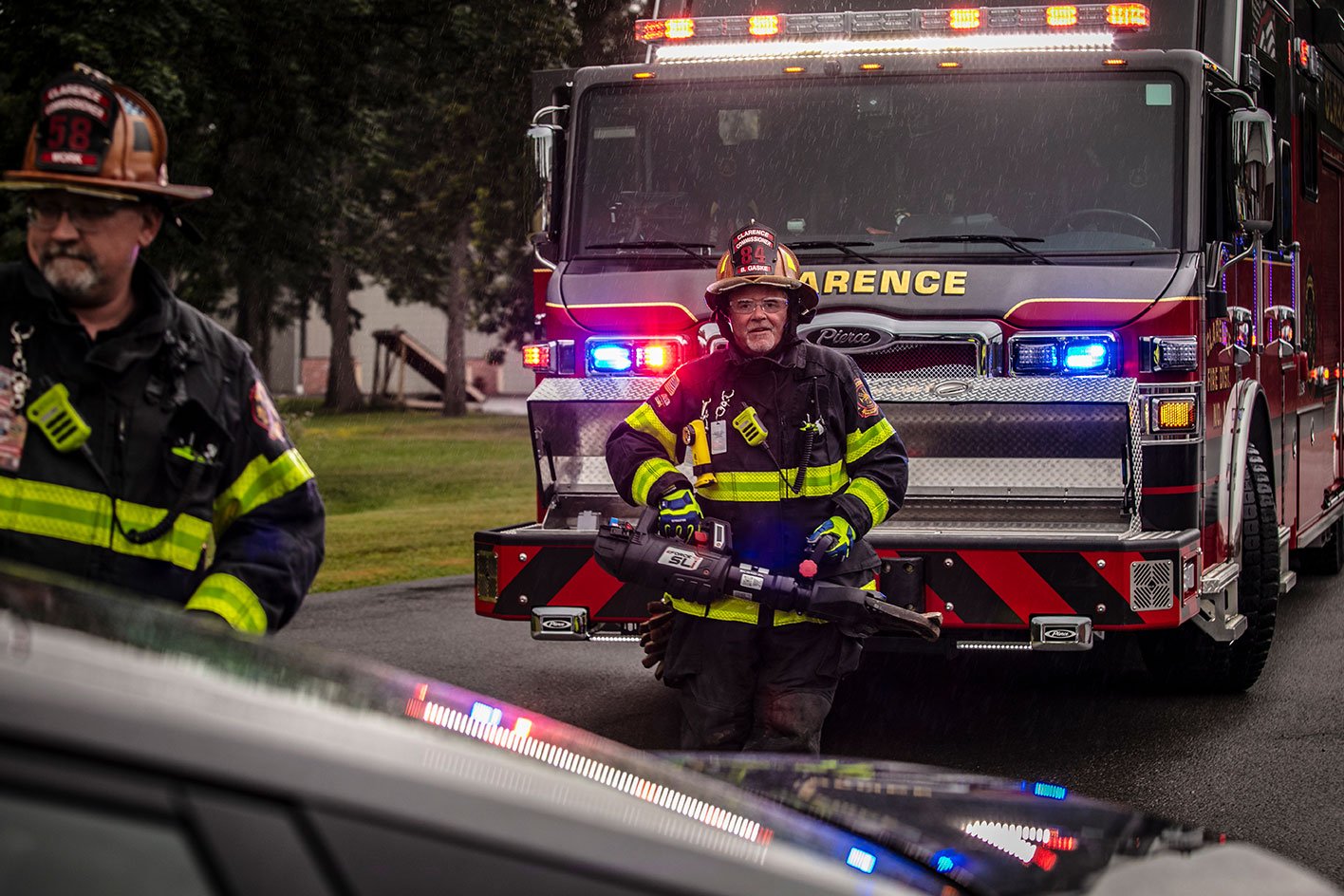 Two firefighters in turnout gear walking in front of a Heavy-Duty Rescue fire truck with extrication equipment on a rainy day.