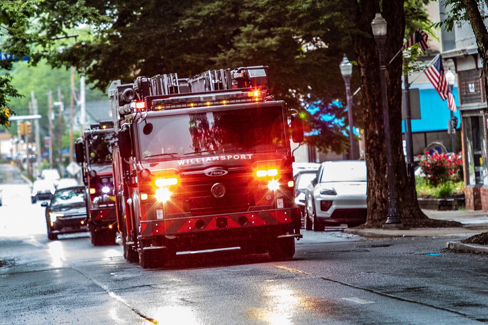 An aerial fire truck driving on a road on a rainy day with the emergency lights on with trees overhanging.