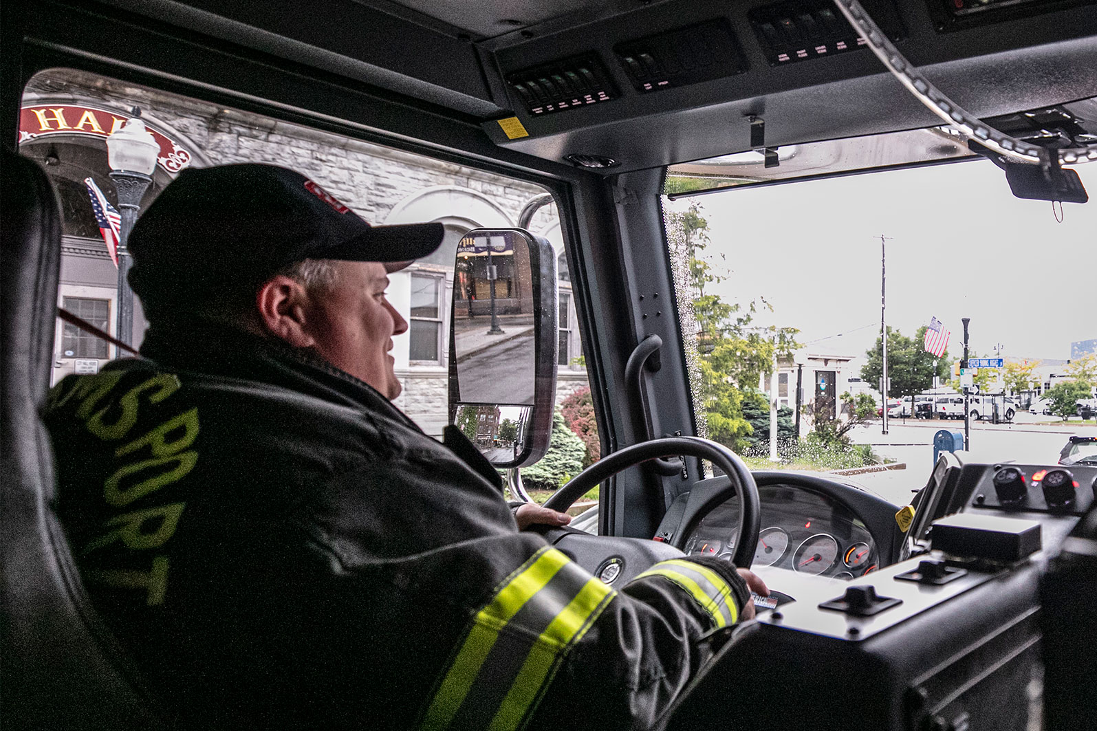 The back of a firefighter in turnout gear driving a fire truck on a suburban road.