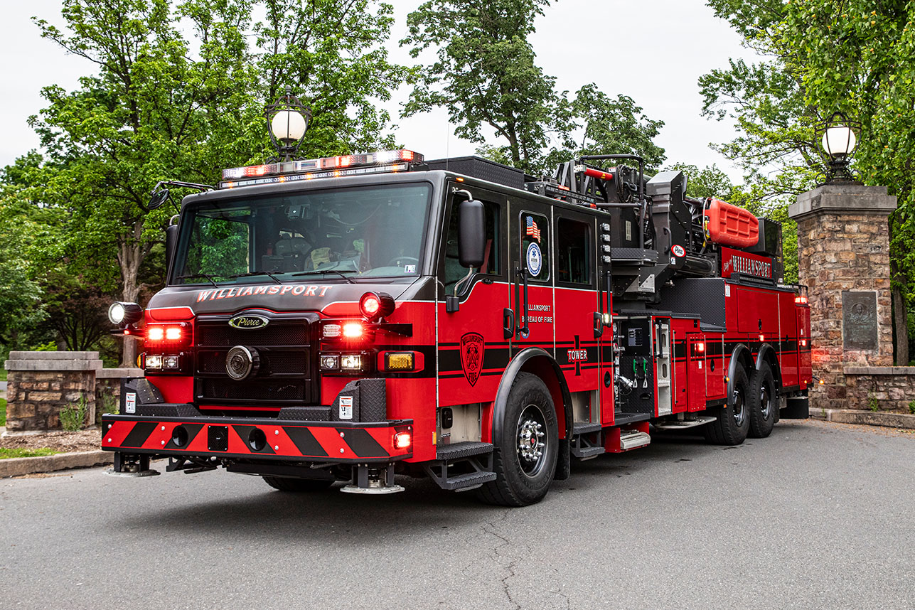 The front driver's side of a Velocity 100' Ascendant Aerial Tower with the lights on driving through a brick entrance.