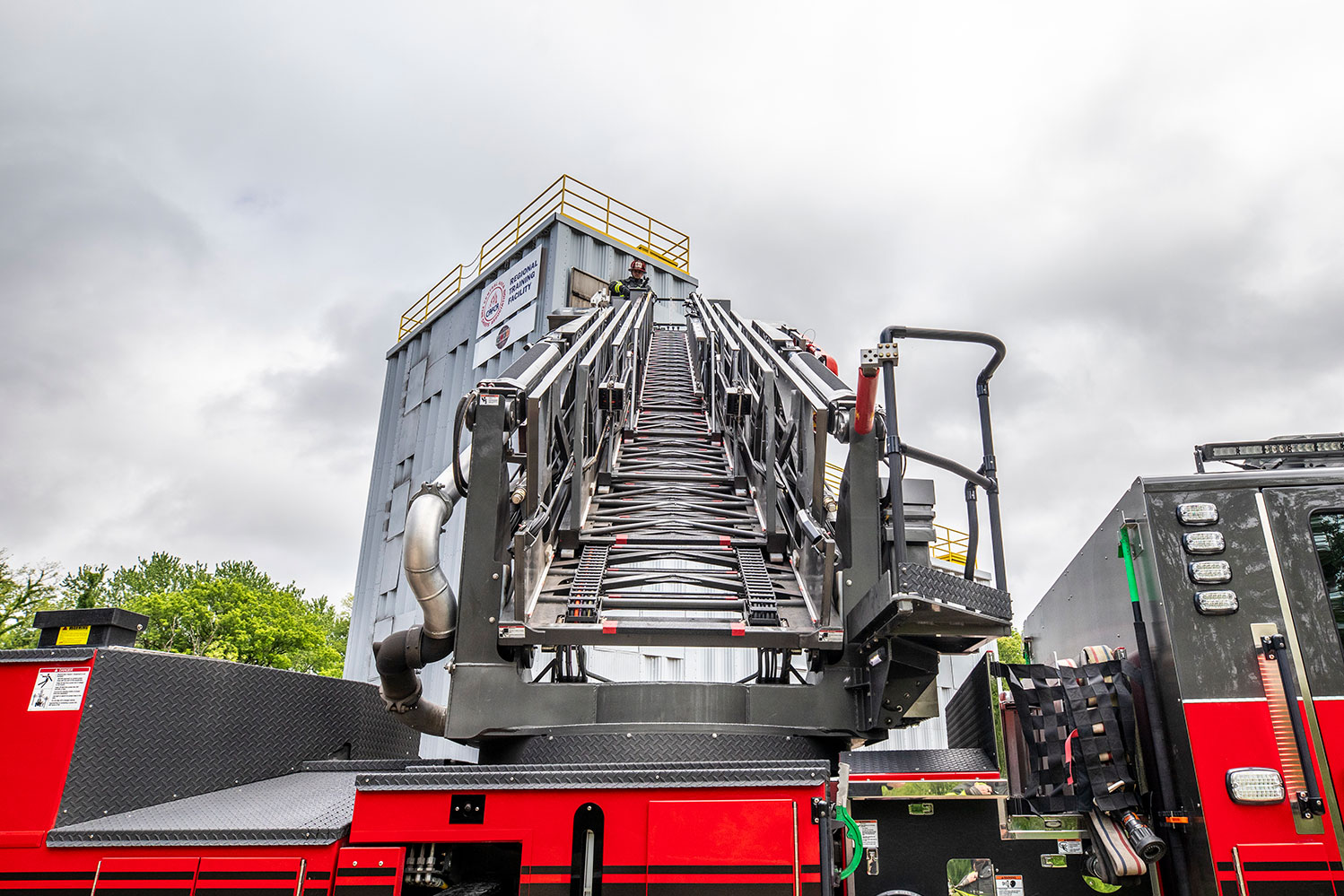 The officer's side of a Mid-Mount fire truck with the aerial extended up towards a firefighter training building.