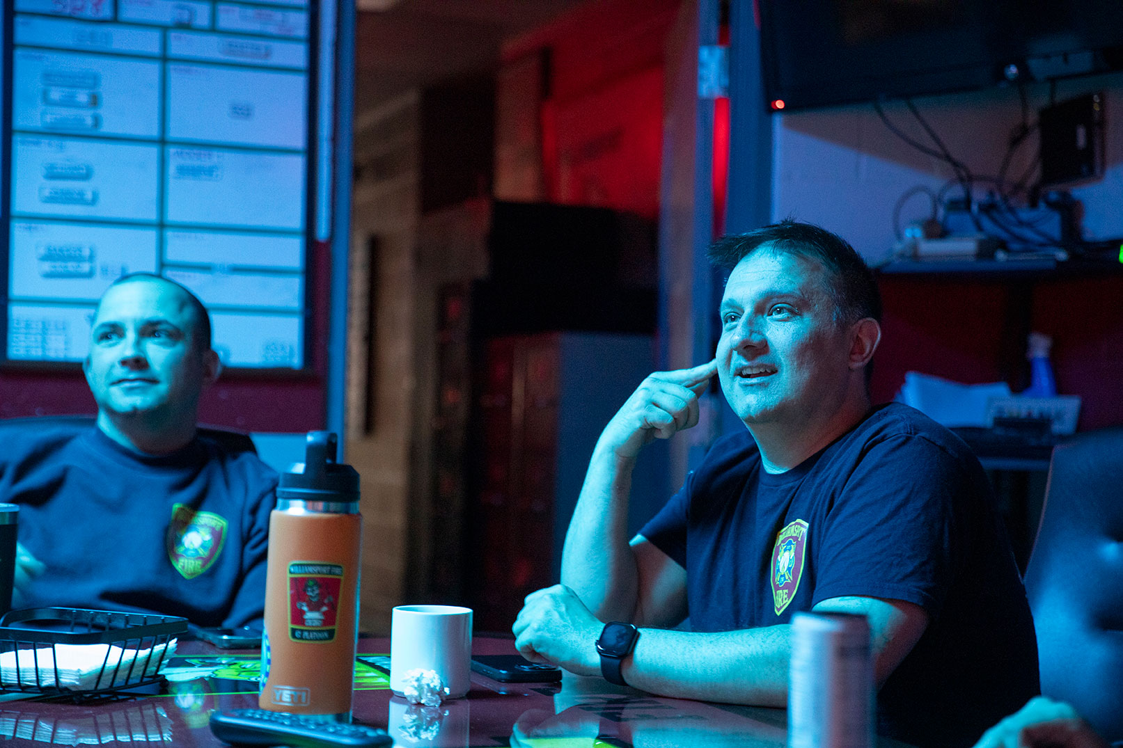 Two firefighters sitting in a fire station looking at a screen with water bottles and coffee mugs on the table.