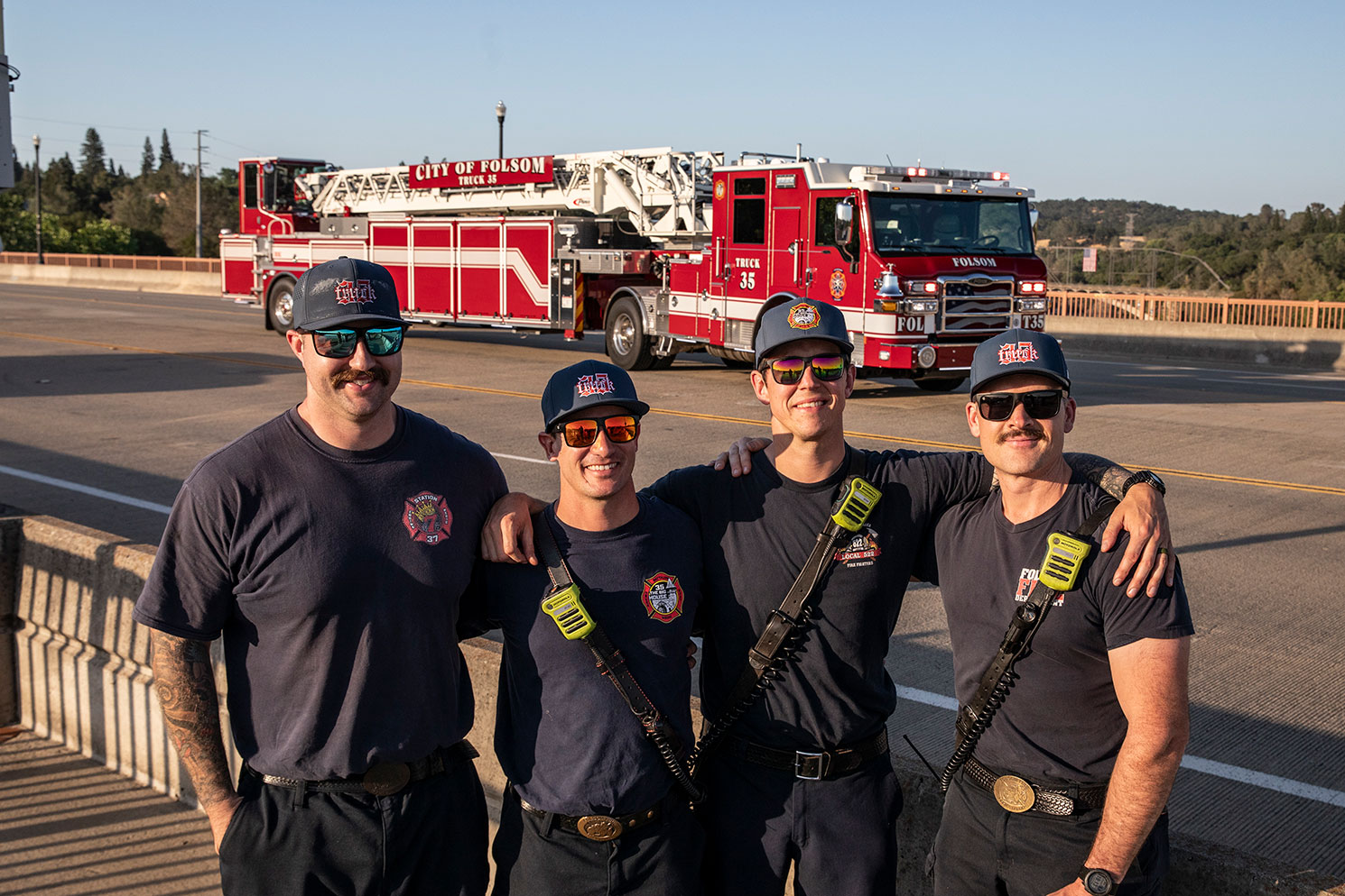 Four firefighters posed together in front of a Tractor Drawn Aerial parked on the road with a rural background.