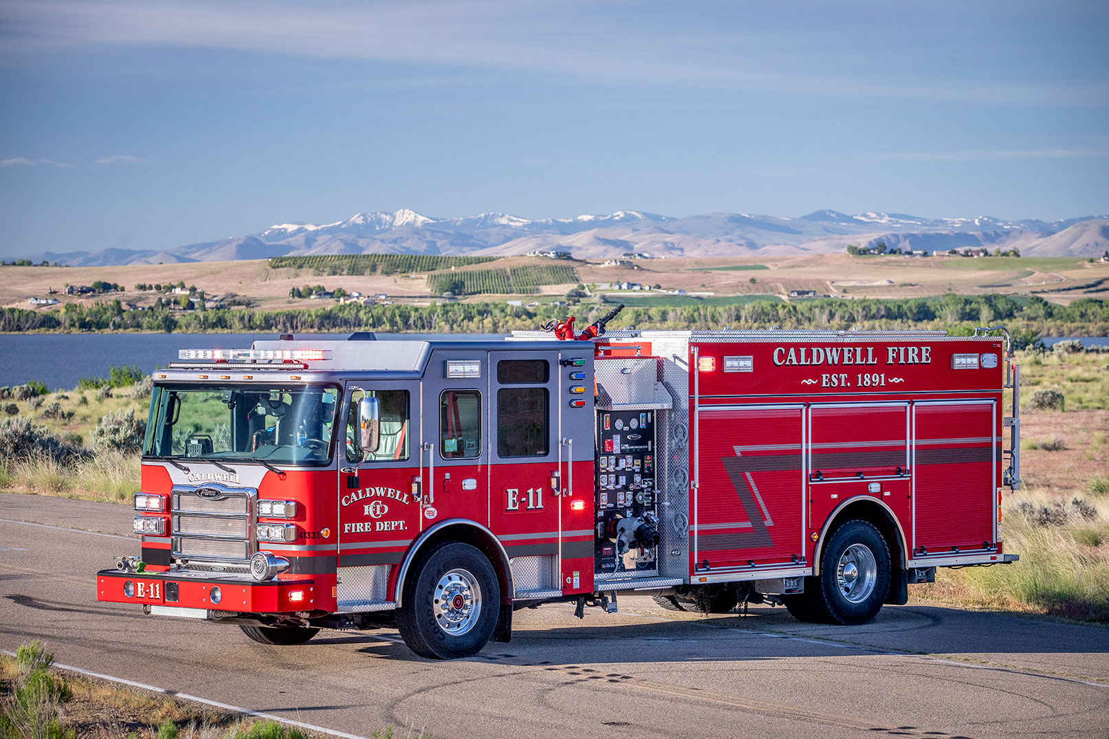 The driver's side of an Enforcer Heavy-Duty Rescue Pumper with the lights on parked on a road in front of a lake and mountains.