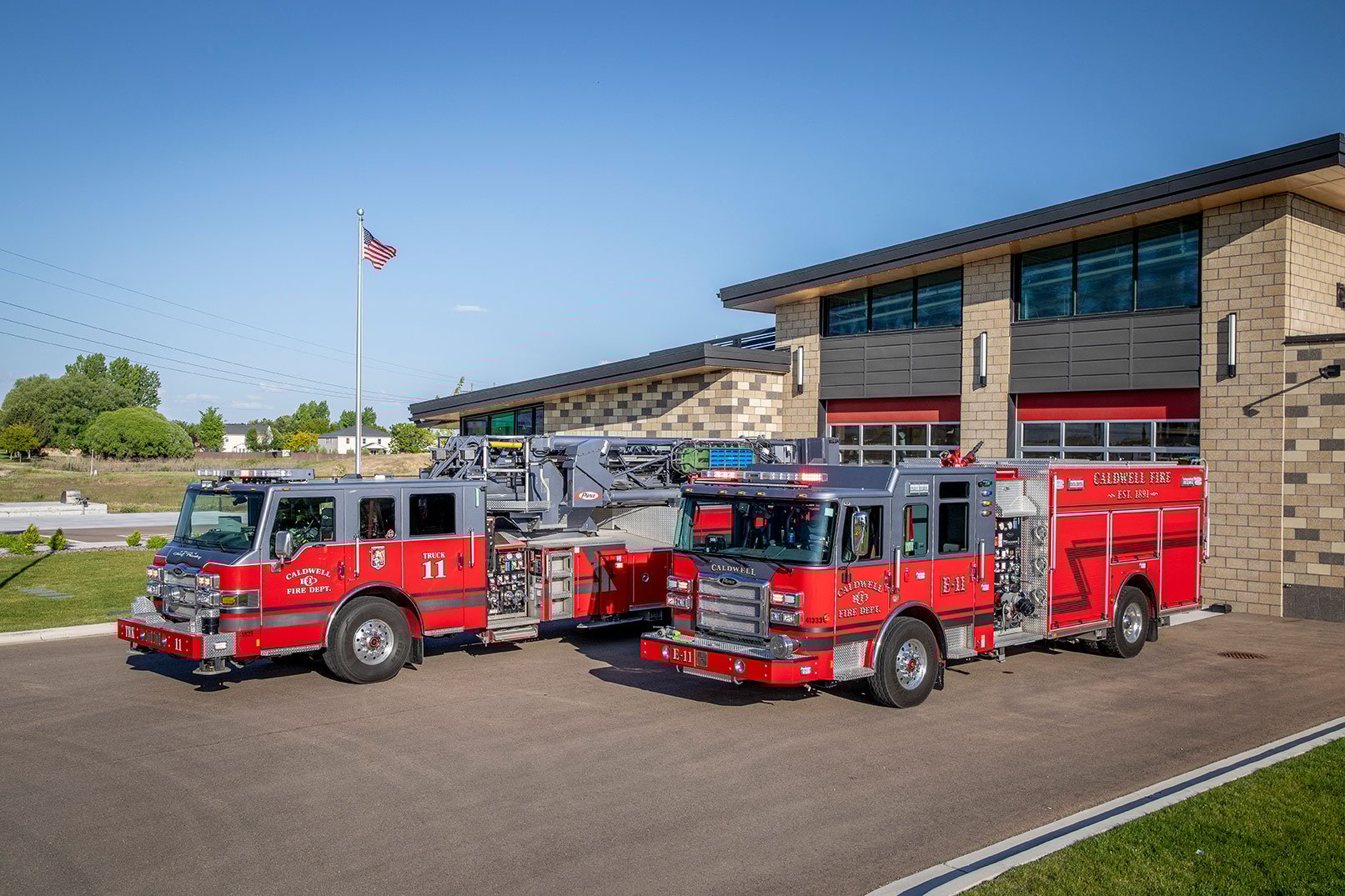 The front driver's side of an Enforcer Heavy-Duty Rescue Pumper and an Ascendant Mid-Mount Tower parked in front of a fire station.