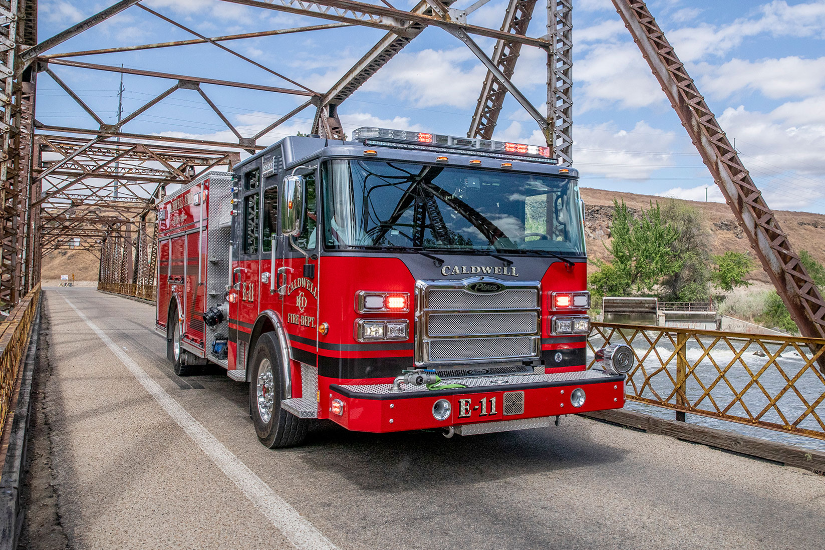 The front officer's side of an Enforcer Heavy-Duty Rescue Pumper driving on a bridge over a dam with the lights on.